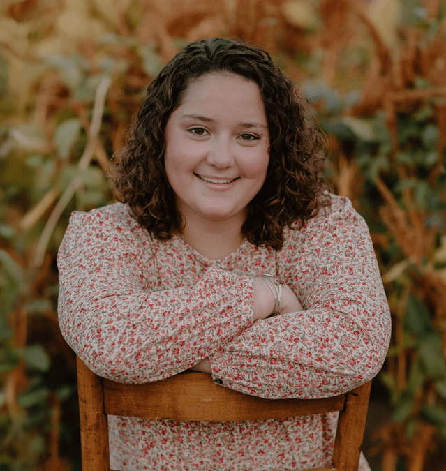 A woman smiling while sitting on a chair, with greenery behind her.